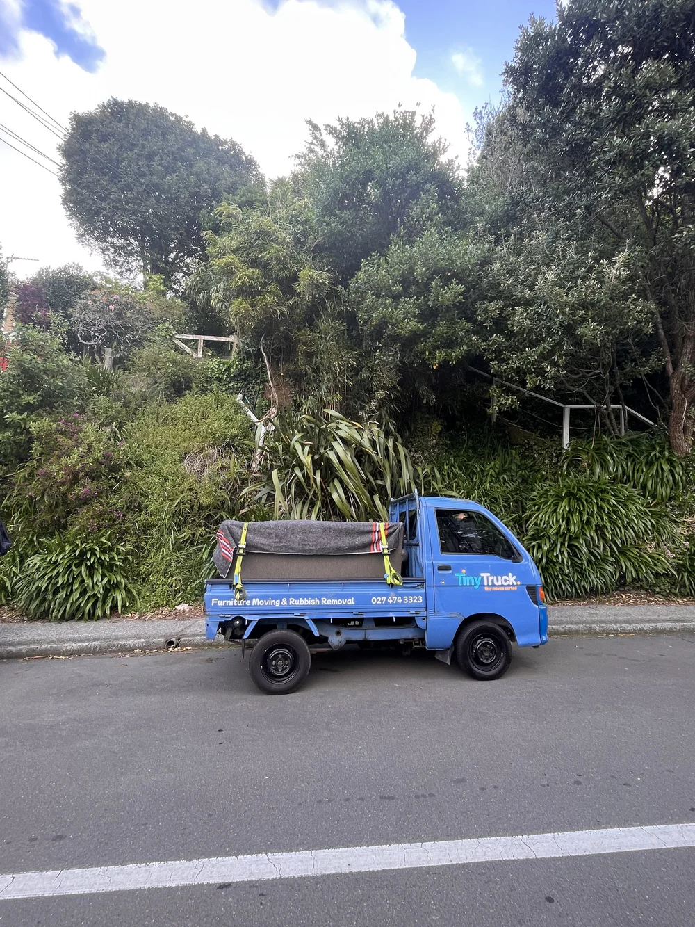 Tiny Truck parked on a bush-lined street with a blanket-covered couch strapped on the back