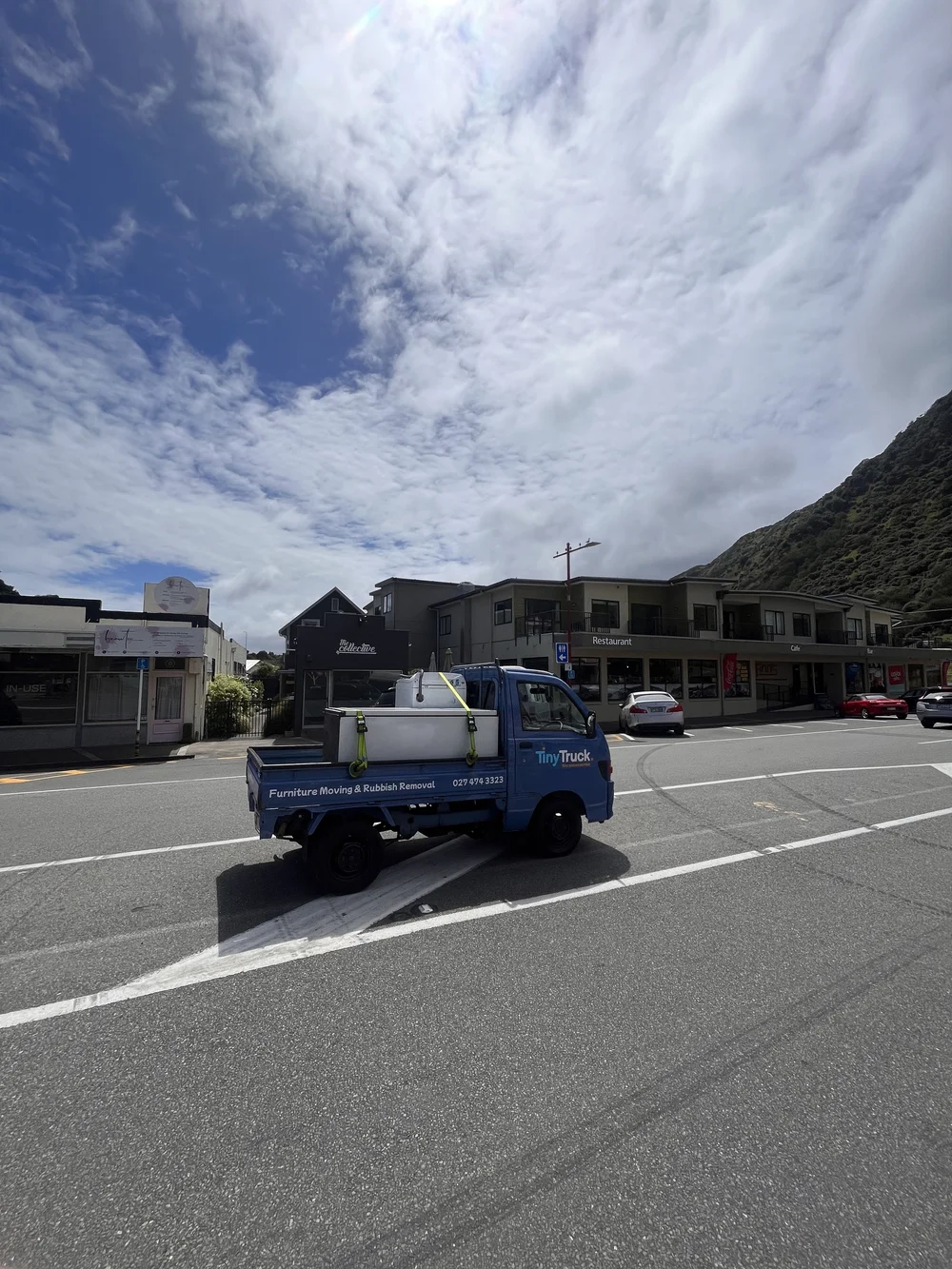 Tiny Truck carrying a white chest freezer through a small-town shopping street