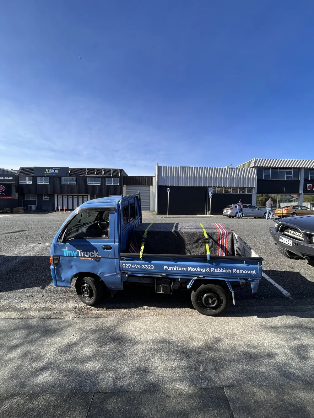 Tiny Truck carrying a blanket-wrapped item strapped down in an industrial car park